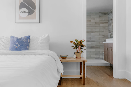 A clean, crisp bedroom with a white comforter and blue accent pillow. There is also a small wooden bedside table with a bowl and plant on it.
