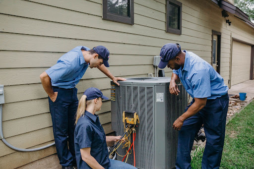 3 HVAC technicians wearing blue uniforms assessing an HVAC unit outside a house
