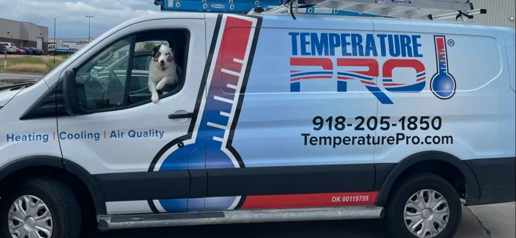 White dog with black and brown spots and blue eyes sticking its head out of a TemperaturePro Tulsa van.