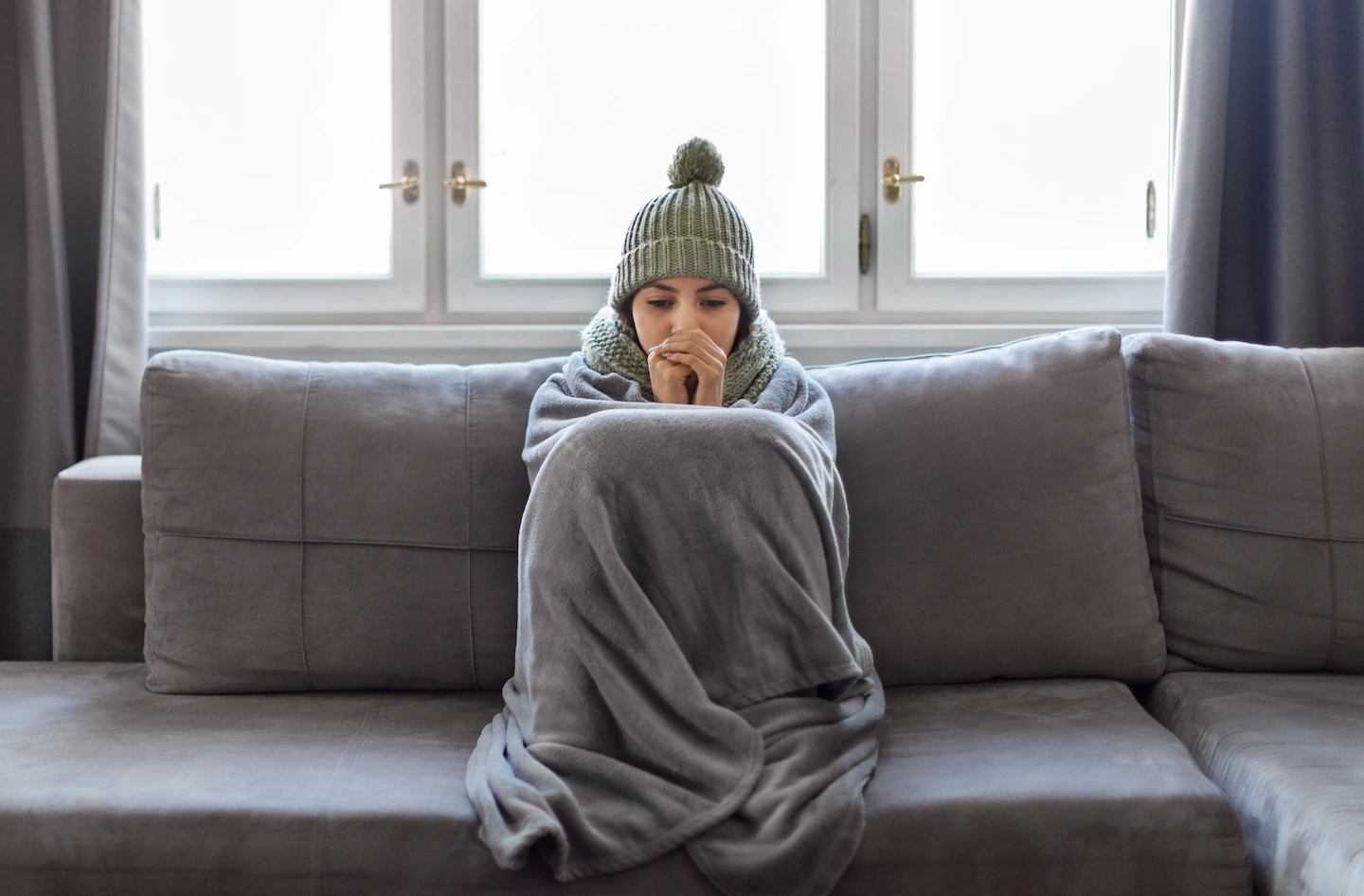 Woman sitting on couch inside with blanket and beanie to stay warm.