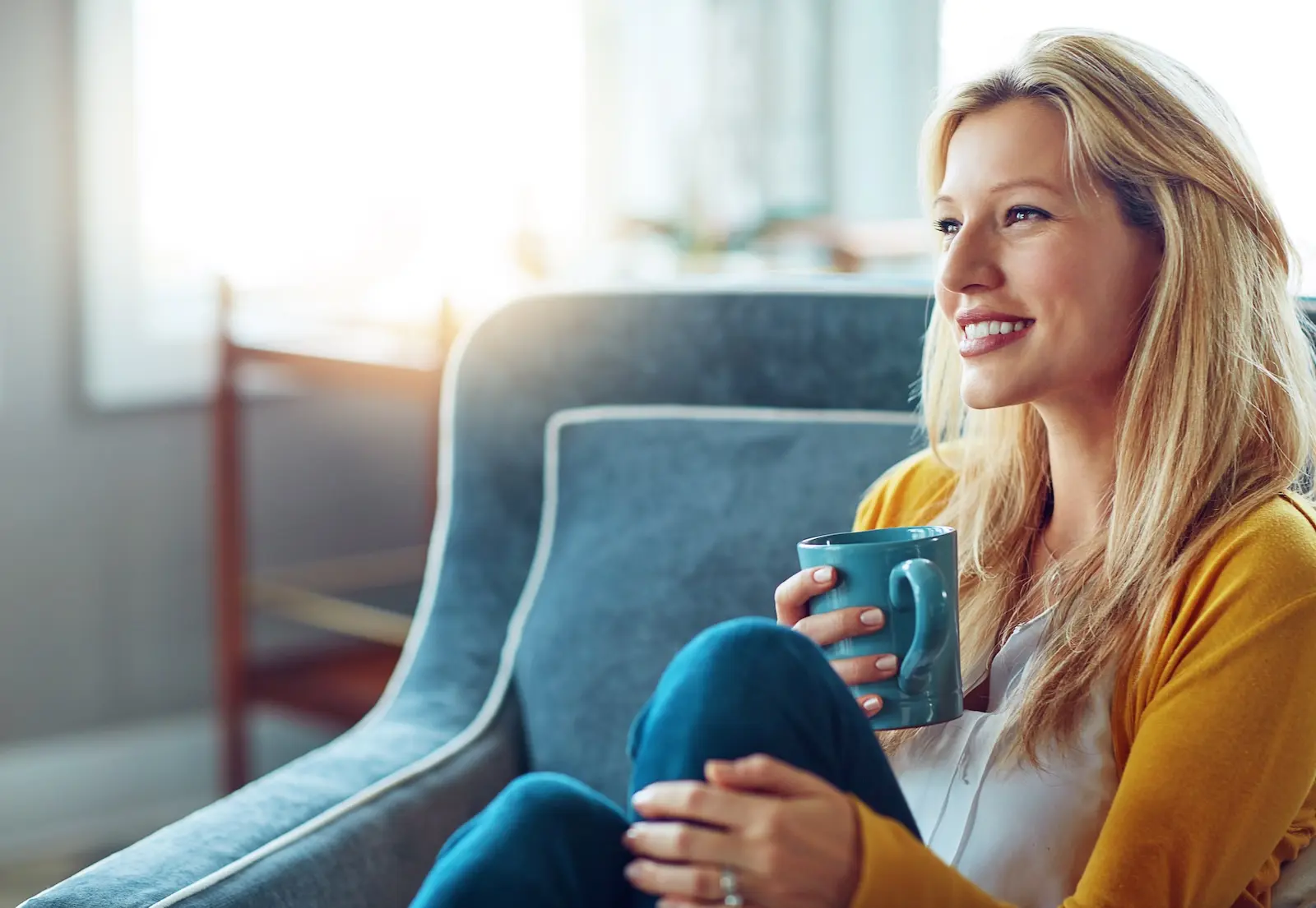 Woman in yellow cardigan holding blue mug sitting on couch while smiling. 