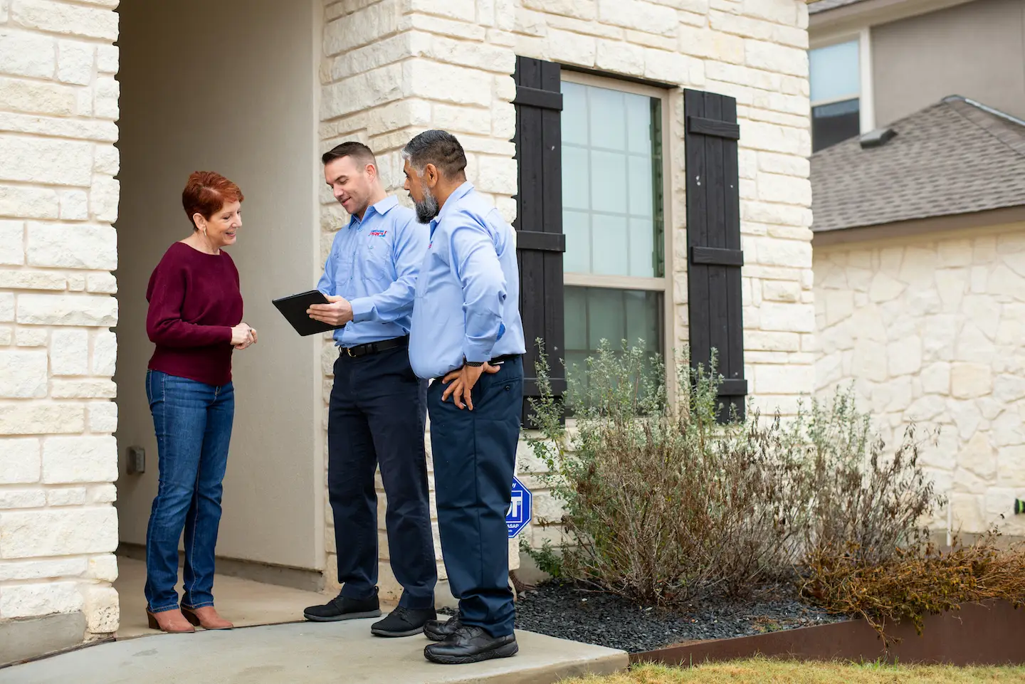 Two HVAC technicians talking to woman in burgundy sweater in front doorway of home.