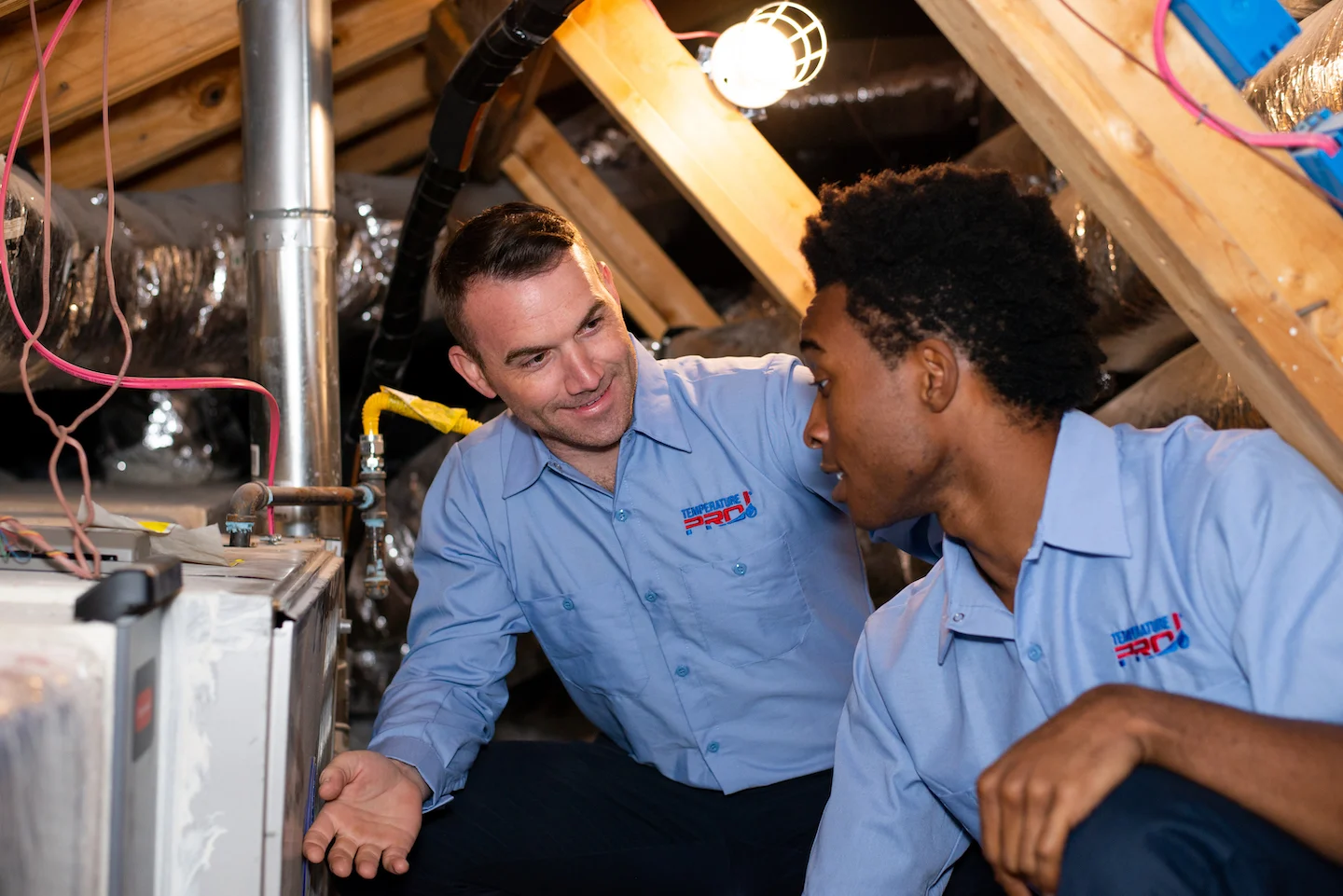 Two technicians in blue collared shirts talking next to a unit in the attic.