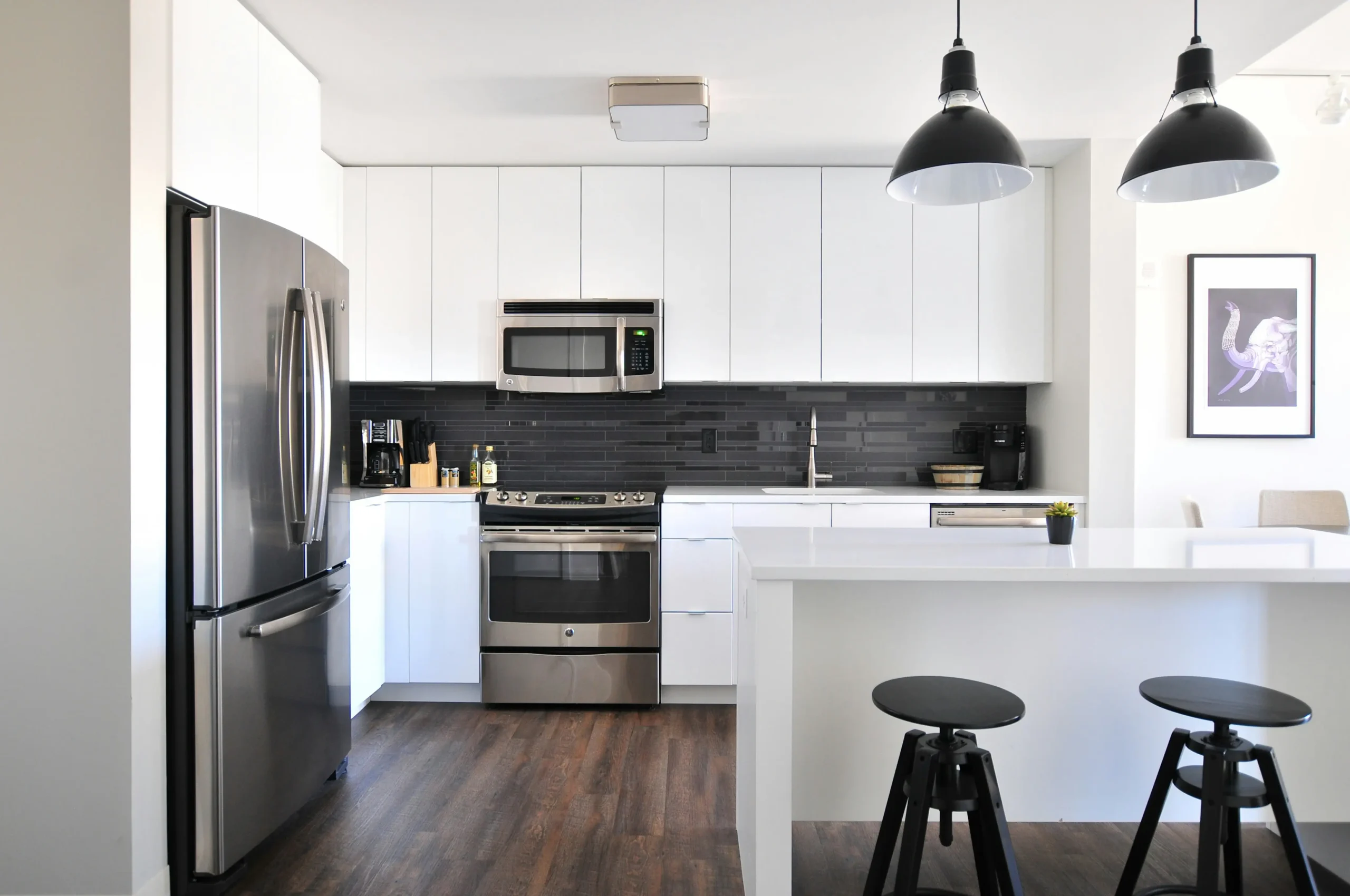 A modern kitchen with a white table and black stools in the front of the photo, with a metallic fridge on the left, and a metallic oven and microwave in the back, attached to white cabinets. 