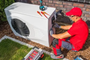 An HVAC technician in a red uniform and cap installs or repairs a white outdoor air conditioning unit next to a brick wall.