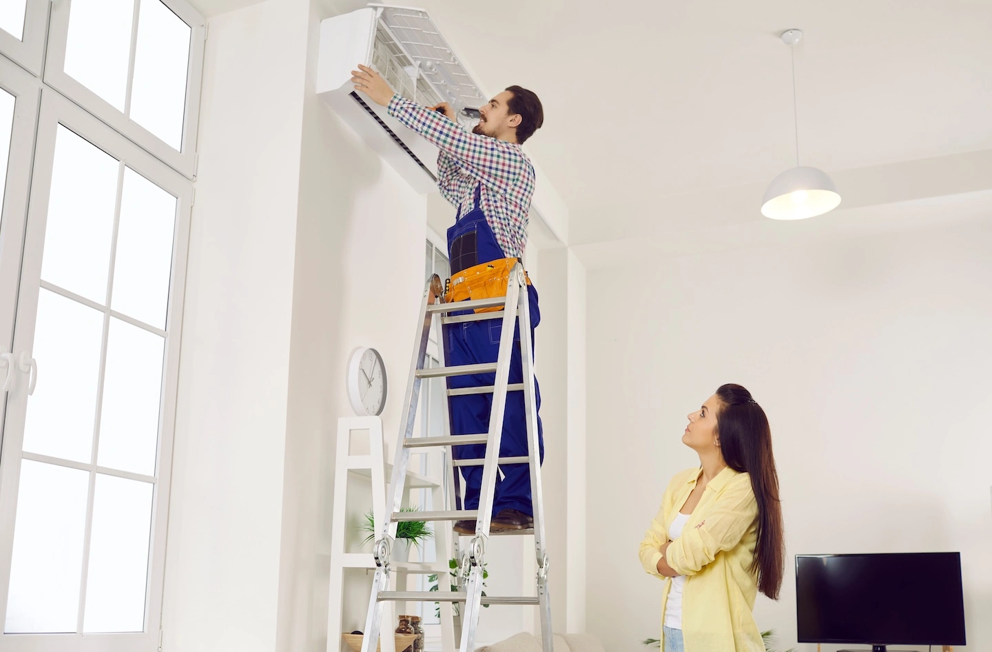 HVAC technician servicing a wall-mounted AC unit while a homeowner looks on.